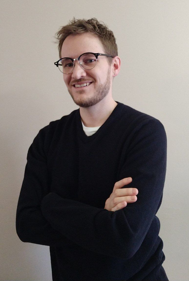 Dylan Hildenbrand poses with his arms crossed. He is a caucasion man wearing glasses, has tossled hair, and a short beard. He wears a black, long-sleeve shirt and has his arms crossed. He is also incredibly handsome. Like, so ridiculously handsome it's not even fair. How many other nerds do you know that look this good?